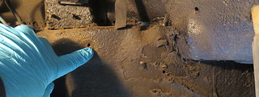 A gloved hand indicating a corrosion perforation through a vehicle frame rail.